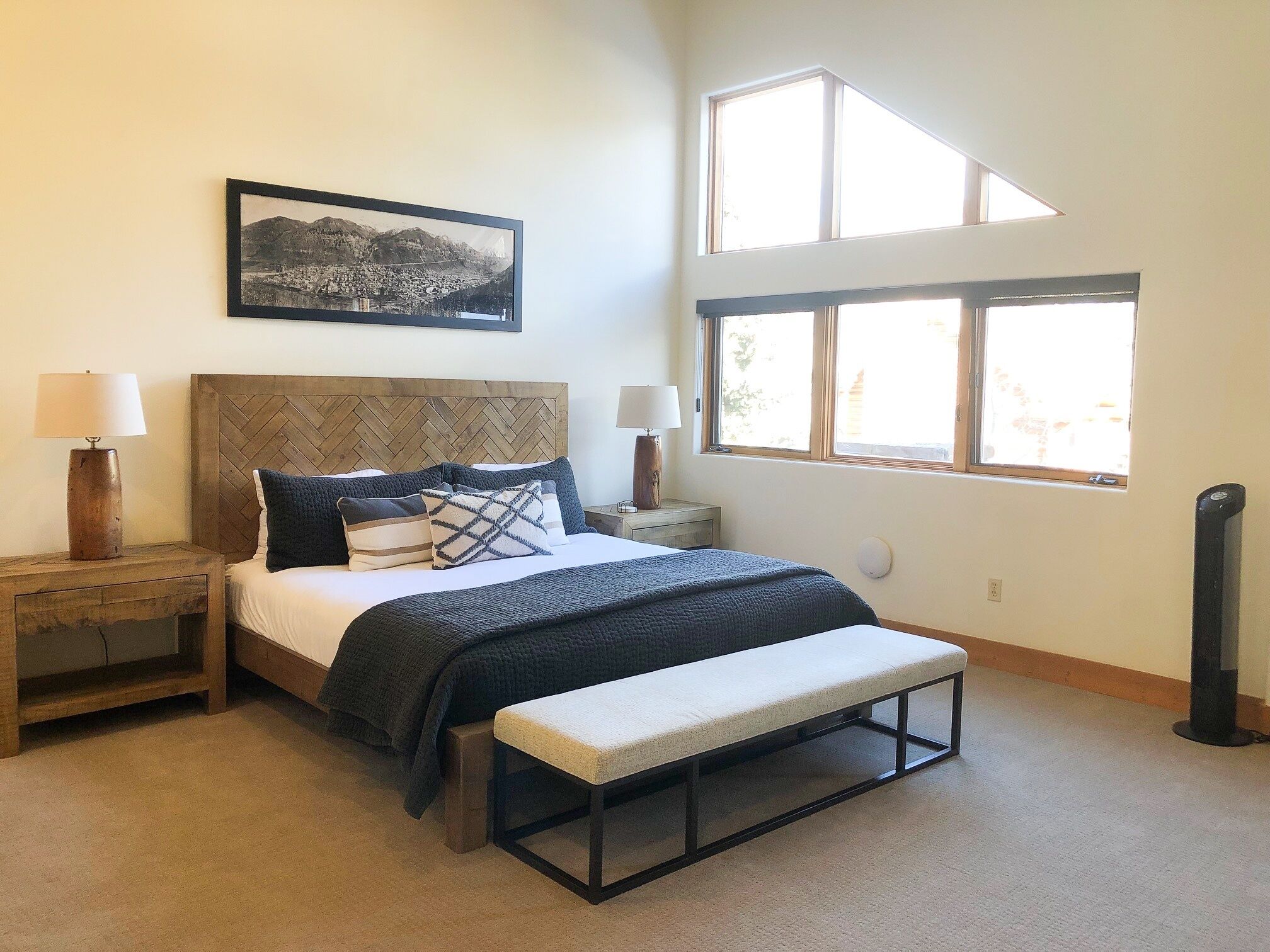 A bedroom with a bed, bedside tables, a bench, a large window, and a picture on the wall.