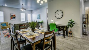 A modern dining area with a wooden table, chairs, and a green accent chair. There is a clock on the wall and a ceiling fan. - 4010 · Comfortable New Home with community pool and golf (Las Cruces)