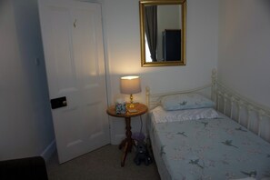 A bedroom with a single bed, a bedside table with a lamp, a mirror above the bed, and a door to the left.
