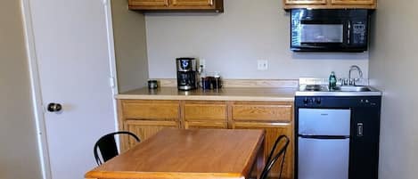 Een kleine keuken met houten kastjes, een magnetron, een koffiezetapparaat, een koelkast en een eettafel met stoelen.