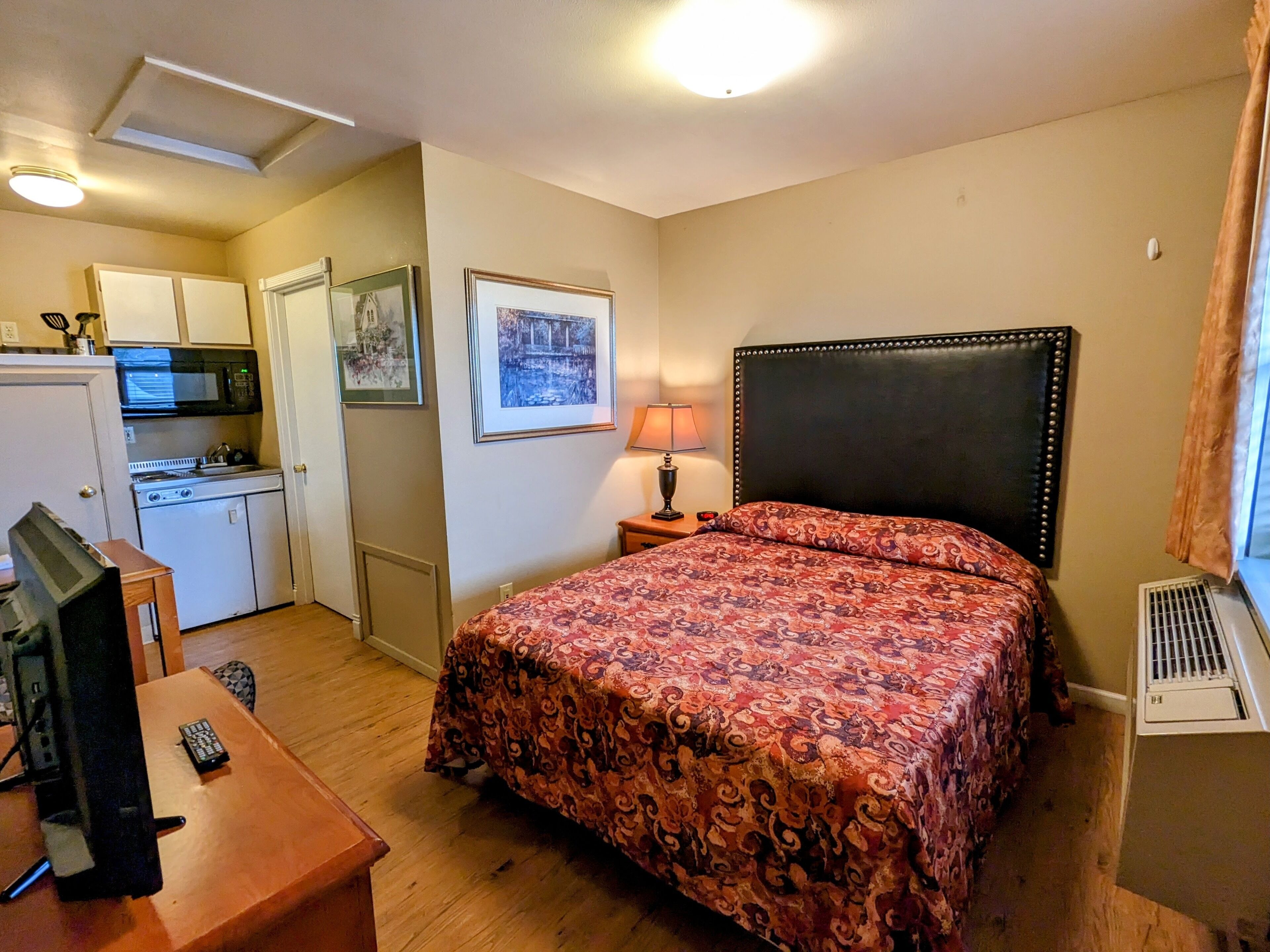 Habitación de hotel pequeña con cama, escritorio, televisión, kitchenette y un cuadro en la pared.