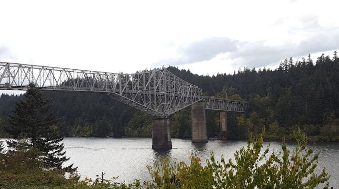 A steel truss bridge over a body of water with forested hills in the background.