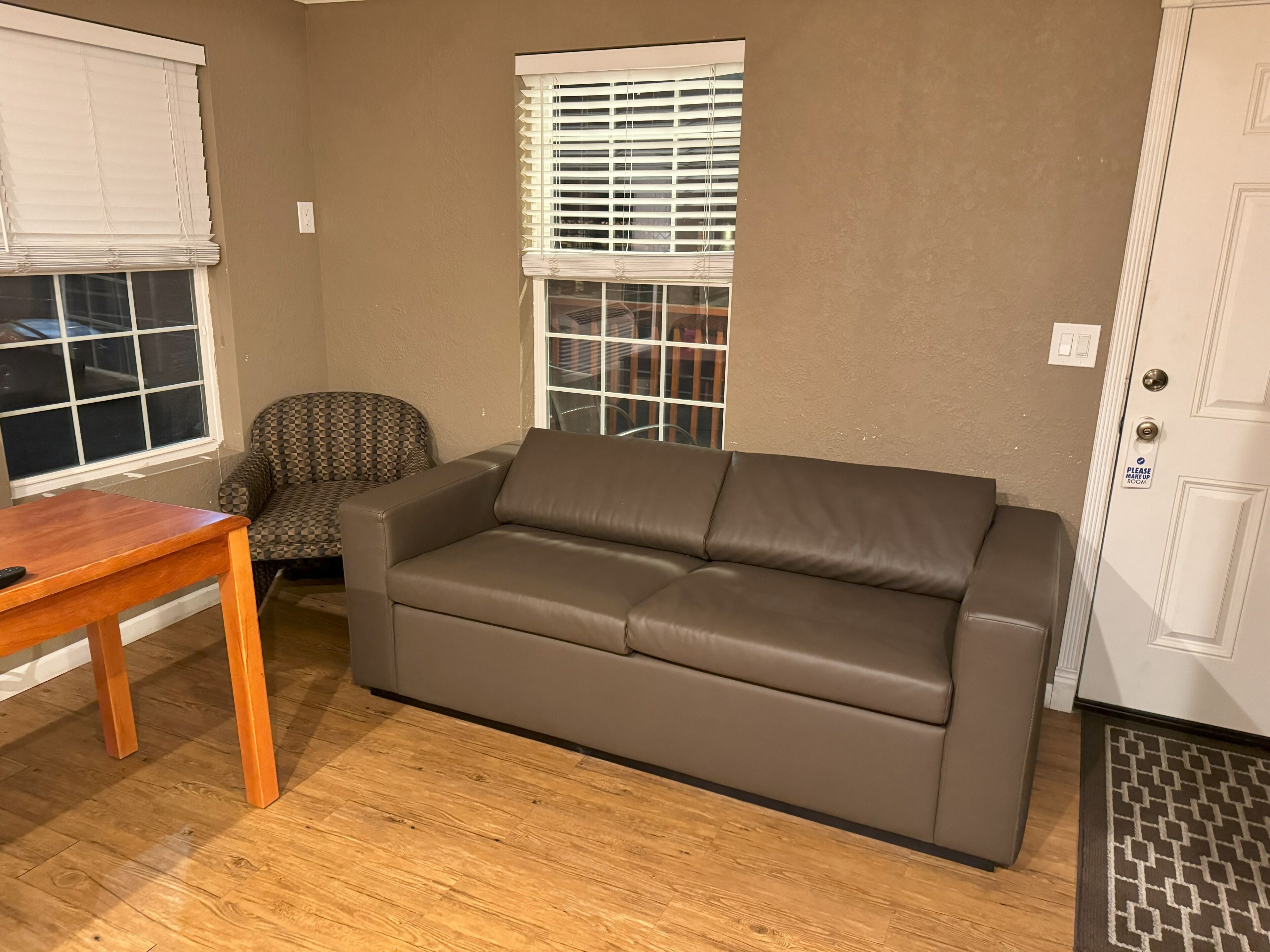 A living room with a brown leather sofa, a patterned armchair, a wooden coffee table, and a door with a 'Please Ask Up' sign.