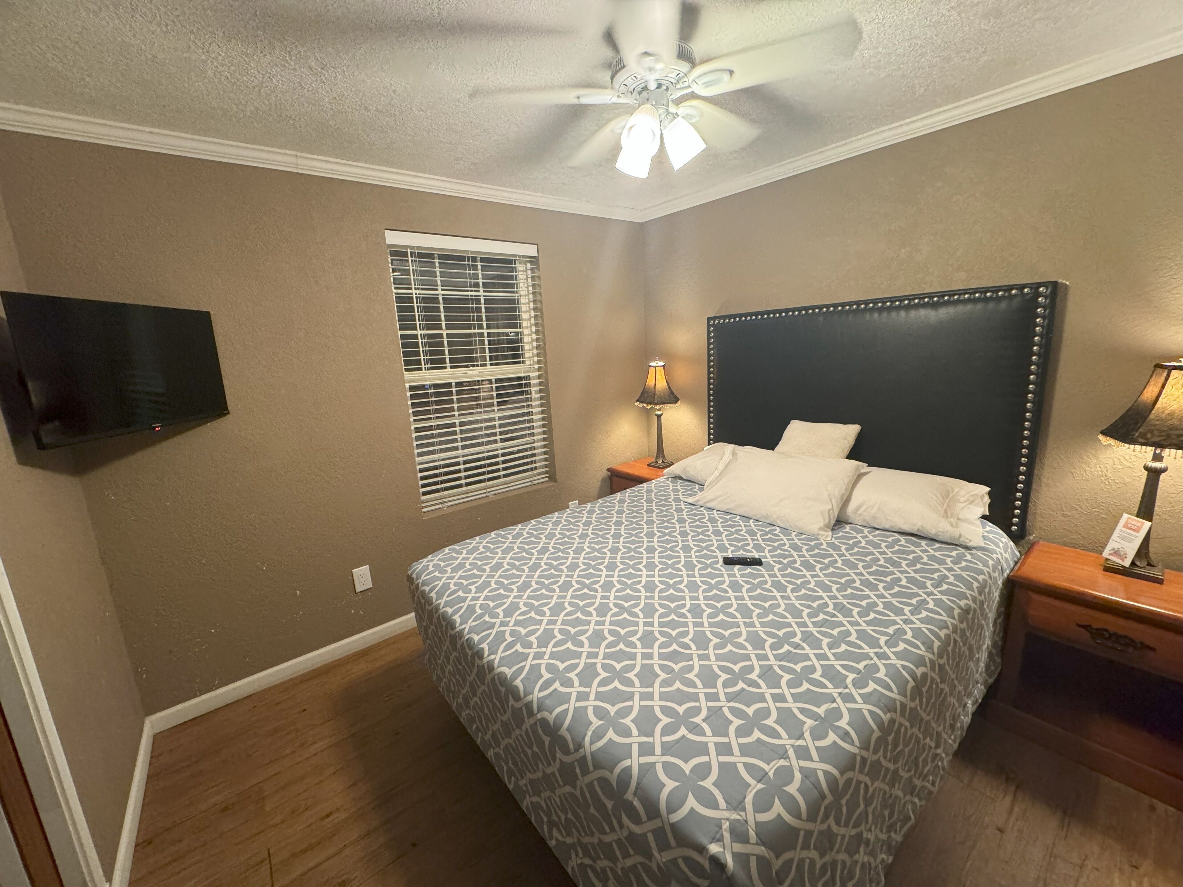 Une chambre à coucher comprenant un lit, deux tables de chevet, un ventilateur de plafond, un téléviseur fixé au mur et une fenêtre avec des stores.