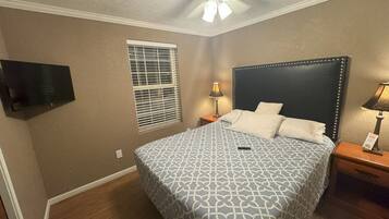 Une chambre à coucher comprenant un lit, deux tables de chevet, un ventilateur de plafond, un téléviseur fixé au mur et une fenêtre avec des stores.