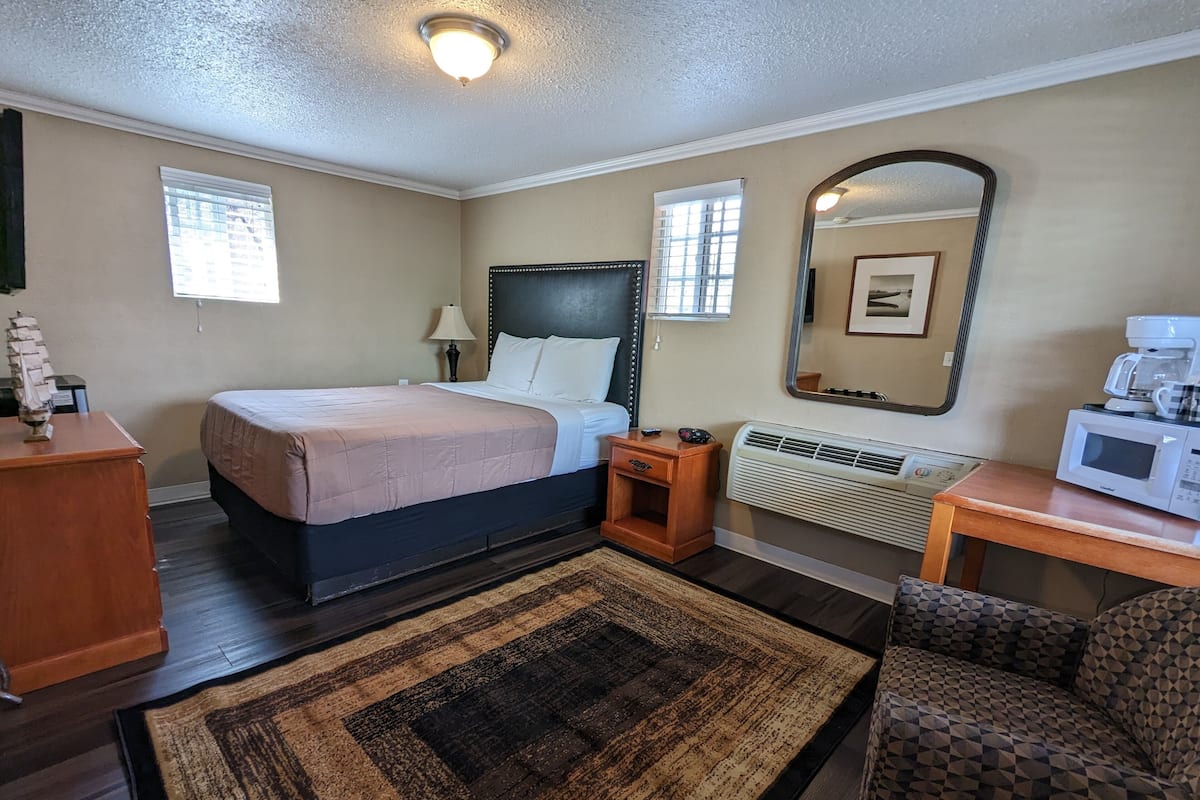 Ein Hotelzimmer mit Bett, Nachttisch, Lampe, Heizung, Mikrowelle und einem Sessel.