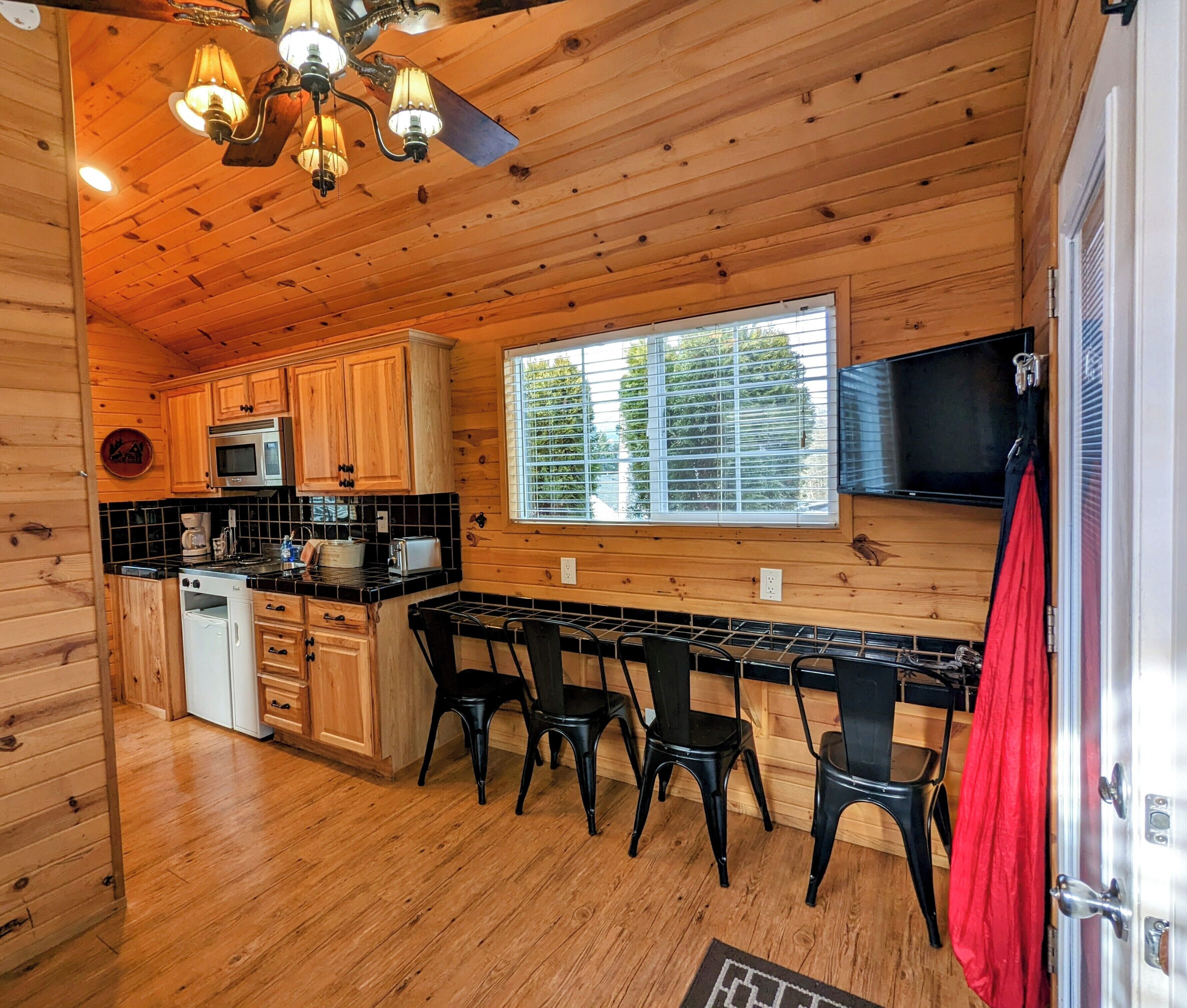 A cabin-style kitchen with wooden walls, a black countertop, and a window overlooking greenery.