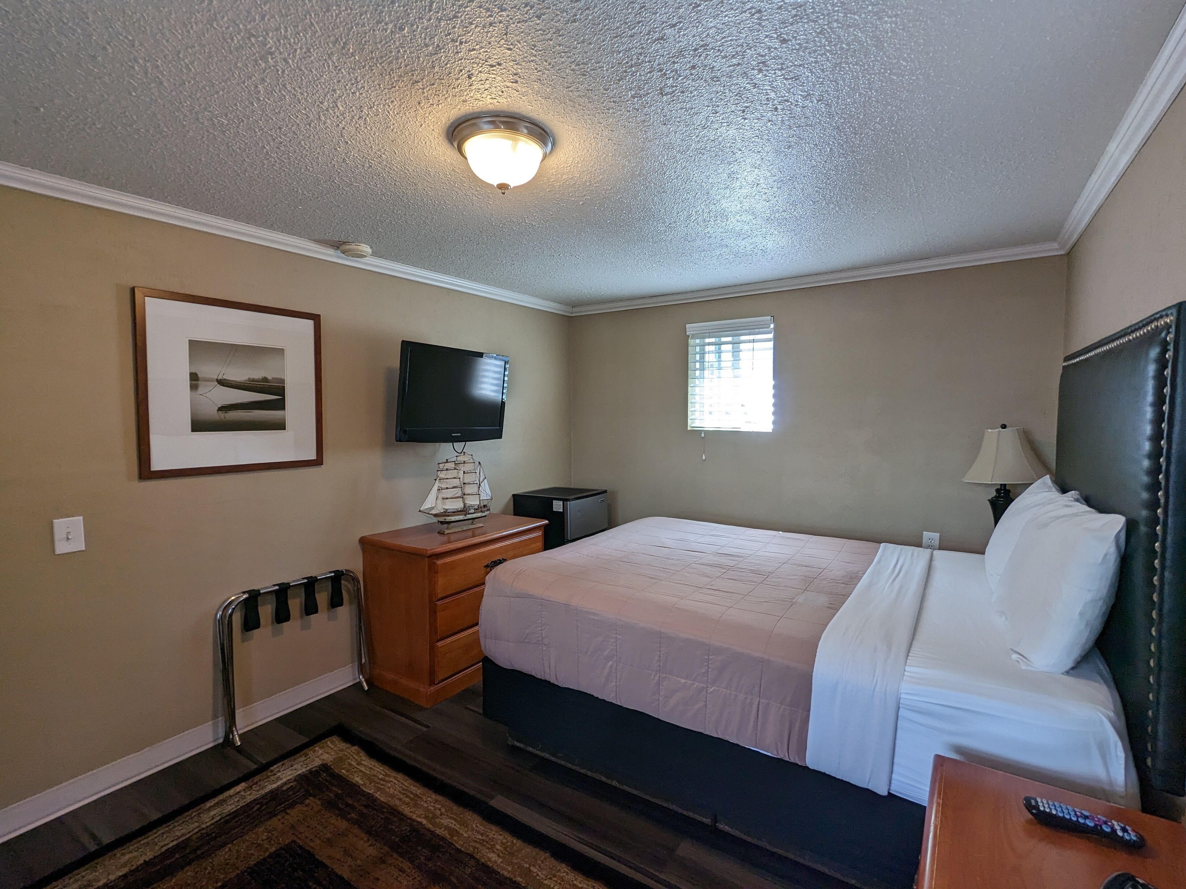 Una habitación de hotel con una cama, un televisor montado en la pared, una mesita de noche con una lámpara, un cuadro enmarcado en la pared y una ventana con persianas.