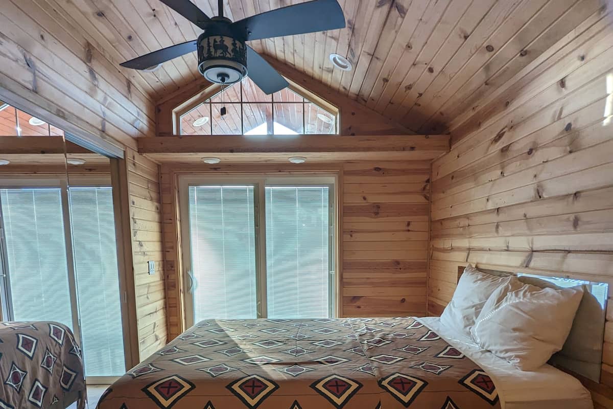 Ein Schlafzimmer einer Holzhütte mit Bett, Deckenventilator und großen Fenstern.