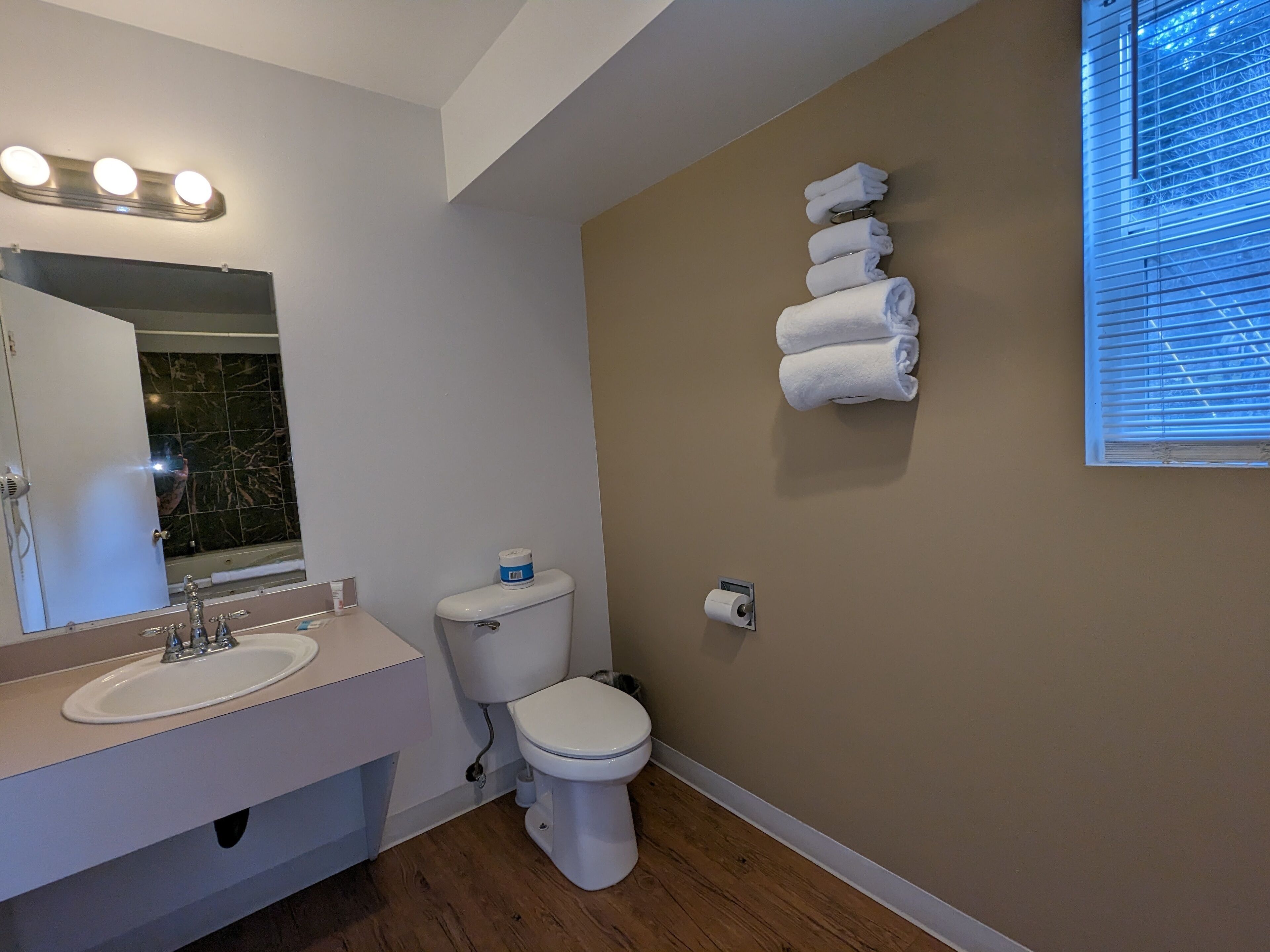A bathroom with a white sink, mirror, and toilet. There are towels hanging on the wall and a window with blinds.