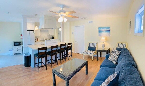 A modern living room with a sofa, a glass coffee table, and a dining area with chairs and a kitchen in the background.