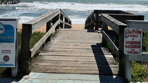 Ein hölzerner Steg führt zum Strand, mit einem Warnschild vor verborgenen Gefahren im Wasser.