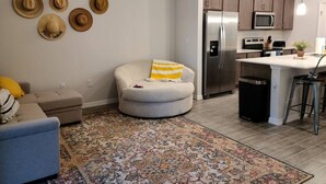 Eine moderne Küche mit Edelstahl-Kühlschrank, Mikrowelle und Herd. Eine kleine Kochinsel mit Barhocker. Das Wohnzimmer verfügt über einen runden Hocker mit gelbem Kissen und einem gemusterten Teppich.