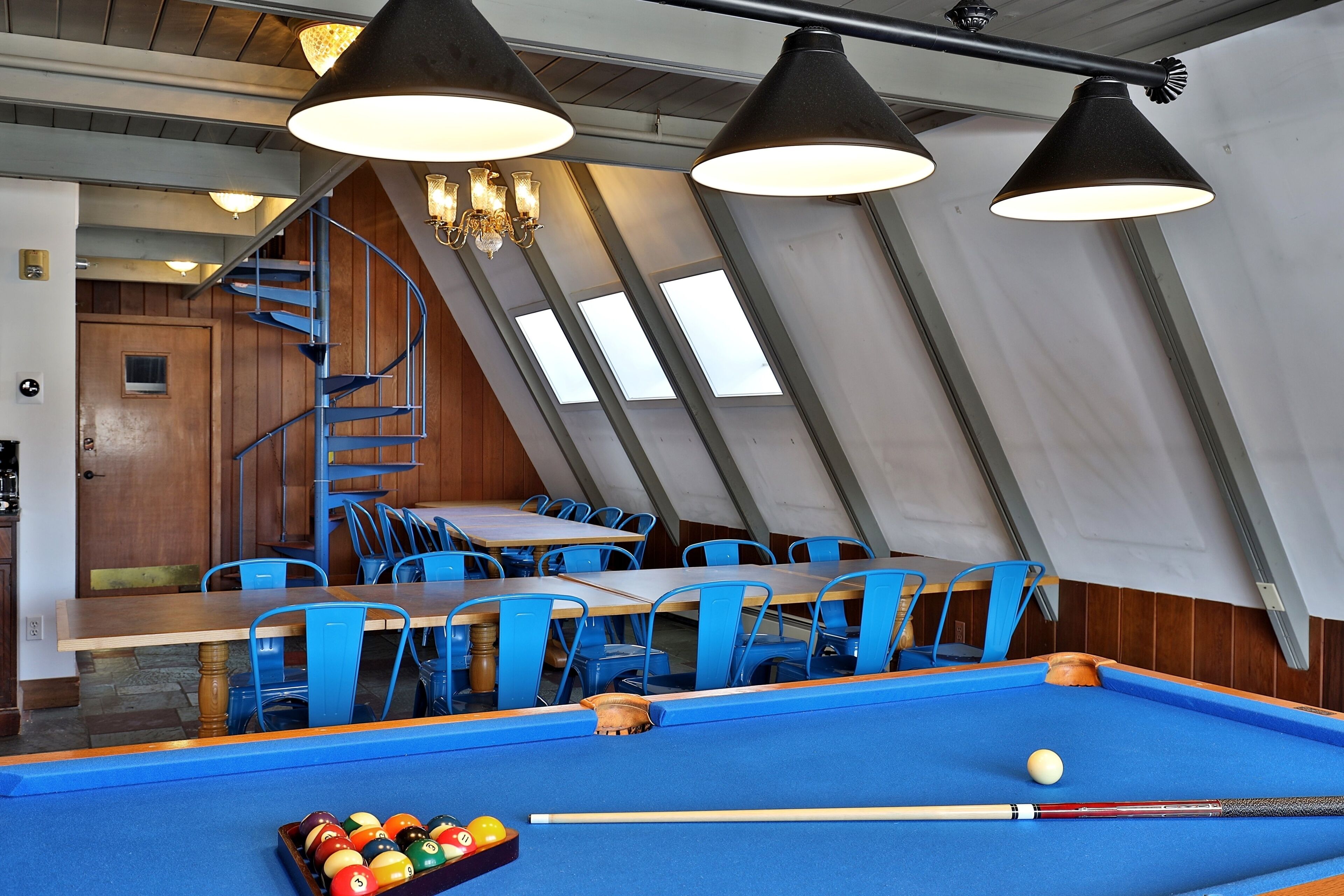 A pool table with a billiard cue and balls, blue chairs, and a staircase leading to a dining area with tables and chairs.