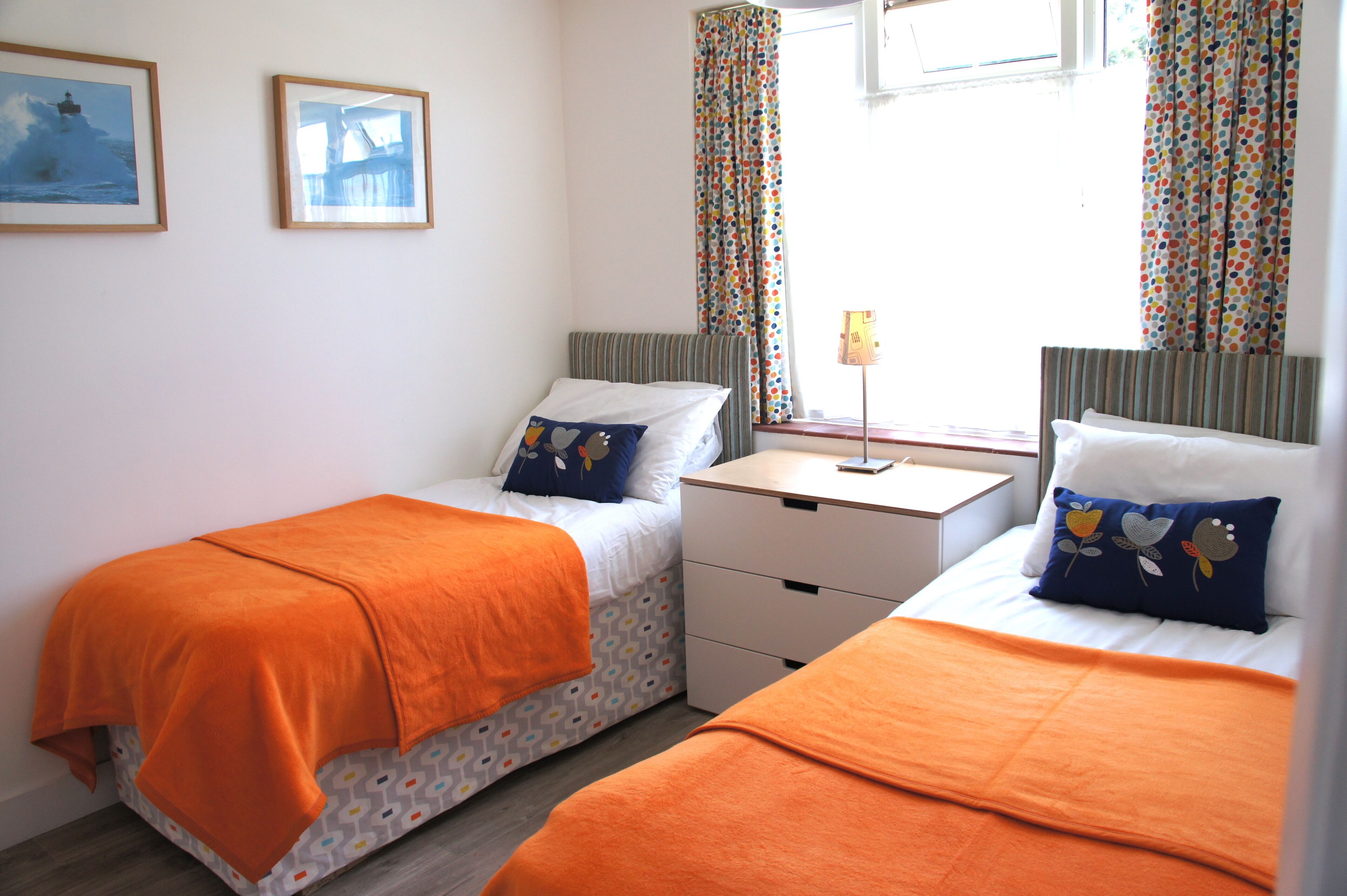 A bedroom with two beds, each with an orange blanket, a bedside table, and a lamp.