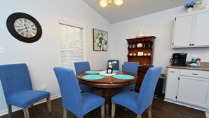 Uma sala de jantar com uma mesa de madeira redonda, rodeada por cadeiras azuis, um relógio de parede, uma janela com persianas e uma área de cozinha com armários e uma cafeteira.