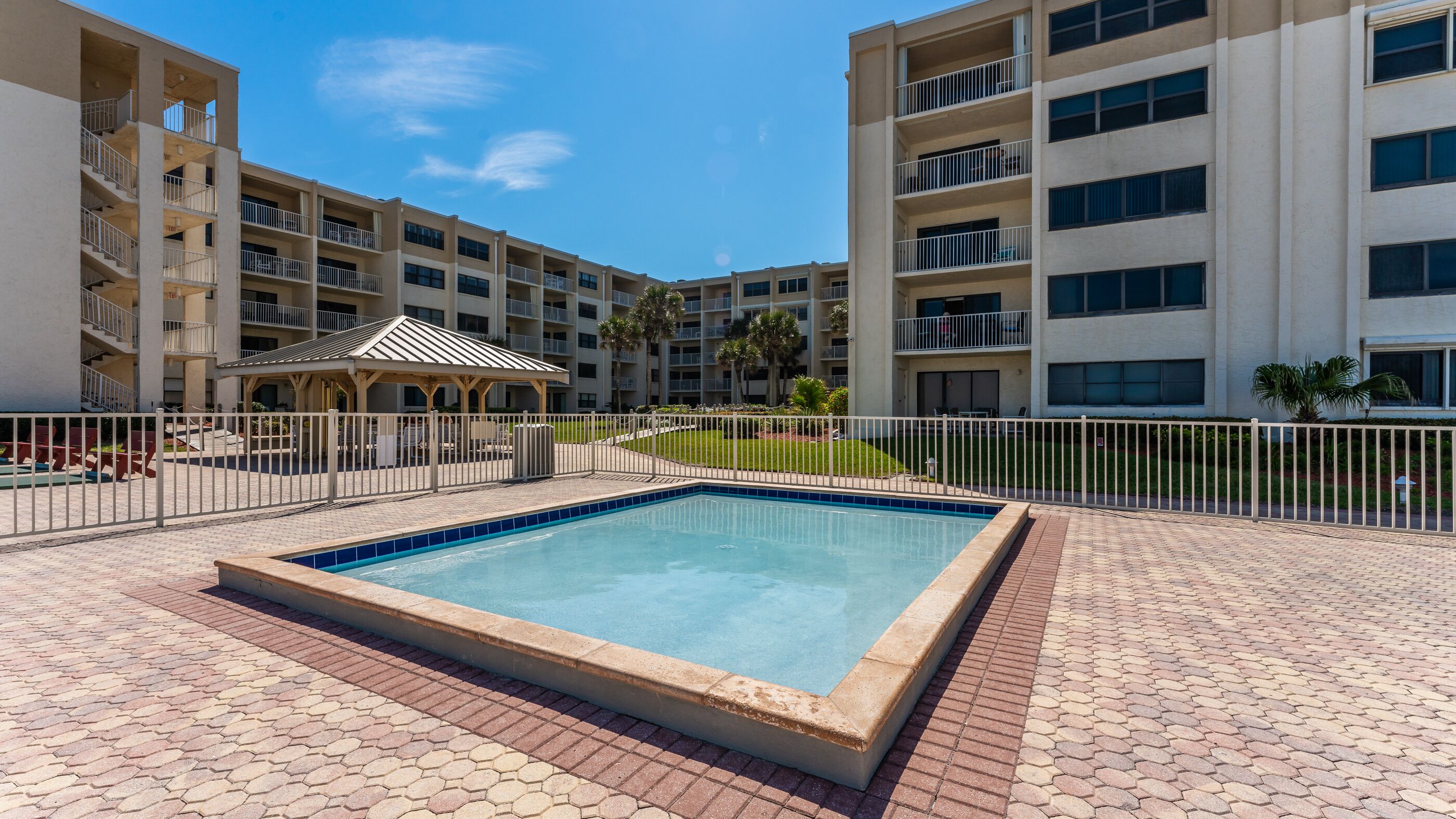 A swimming pool with a tiled deck, surrounded by a fence, and a covered seating area with chairs and tables.