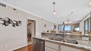 A modern kitchen with a dishwasher, granite countertop, stainless steel sink, and a view of the ocean through large windows.