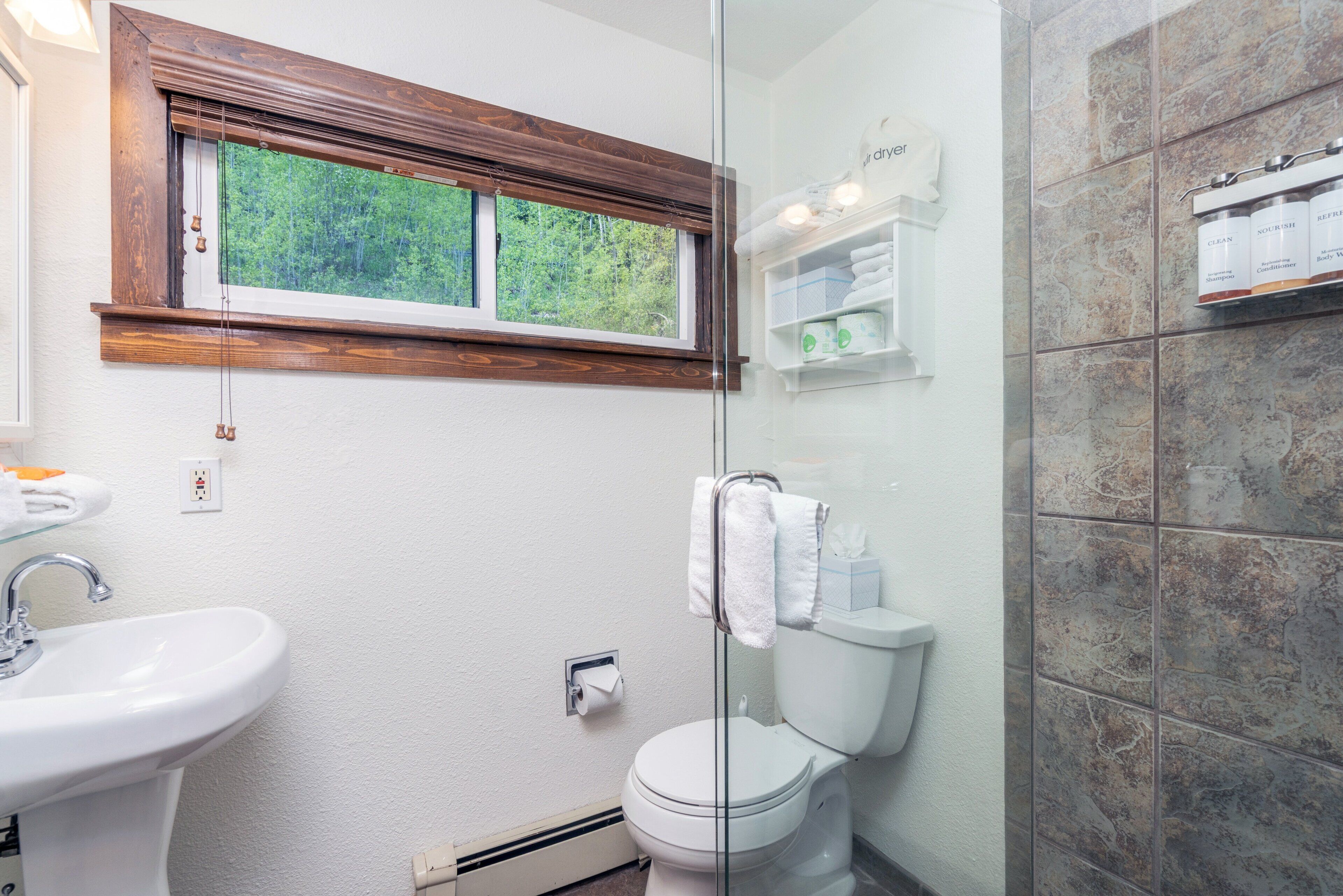 A bathroom with a white sink, a toilet, a window with a wooden frame, and a shower area with tiled walls.