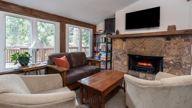 A living room with a stone fireplace, a flat-screen TV, a bookshelf, and a window with a view of trees. - Manitou Lodge 3 Hotel Room by Alpine Lodging Telluride (Telluride)