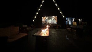 An outdoor seating area with a long wooden table and benches, illuminated by string lights forming a triangular pattern. - The Birch Ridge: American Classic Room #7 - King Suite In Killington. Hot Tub. 1 Bedroom Home (Killington)