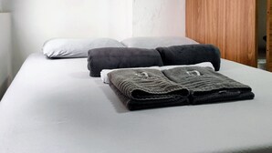 Cama com colchão branco, duas almofadas escuras e um cobertor escuro dobrado. Uma porta de madeira está entreaberta à direita.