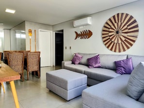 Una sala moderna con un sofá gris, cojines morados, una zona de comedor con sillas de mimbre y un pez decorativo colgado en la pared.