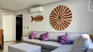 Uma sala de estar moderna com um sofá seccional cinza, duas almofadas roxas, uma peça de arte redonda na parede e um ar condicionado branco.