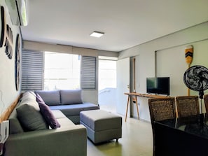Una sala de estar con sofá, reposapiés, televisor, ventilador y una ventana con persianas.