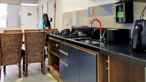 Cozinha moderna com armários escuros, bancada preta e torneira vermelha. Há cadeiras de vime e uma pequena mesa de jantar.
