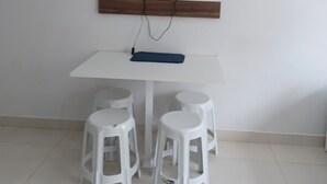 Una mesa blanca con un televisor de pantalla plana montado en un panel de madera encima. Hay tres taburetes de plástico blanco alrededor de la mesa.