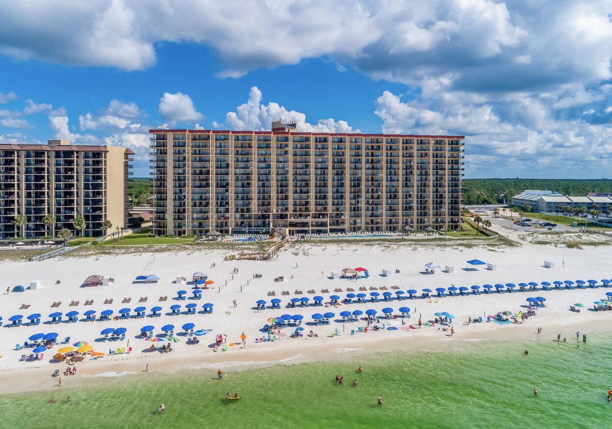 A beachfront hotel with multiple high-rise buildings, a sandy beach with numerous umbrellas, and a clear blue sky.