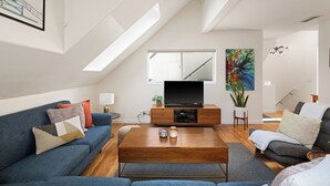 Una sala de estar moderna con sofá, mesa de centro y televisor. Hay un cuadro en la pared y una escalera que sube a un nivel superior.