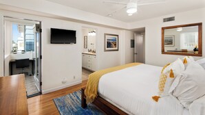 Un dormitorio moderno con cama, escritorio, televisor y baño visible a través de una puerta abierta.