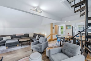 A spacious living area with a sofa set, armchairs, a coffee table, and a dining table with chairs. A staircase leads to an upper level. - Quechee Club Pool & Golf | 4BR Kingswood 10A (Quechee)