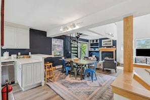 A modern kitchen with white cabinets, a dining area with blue chairs, a fireplace, and a large TV. - Quechee Club Pool & Golf | 4BR Kingswood 10A (Quechee)