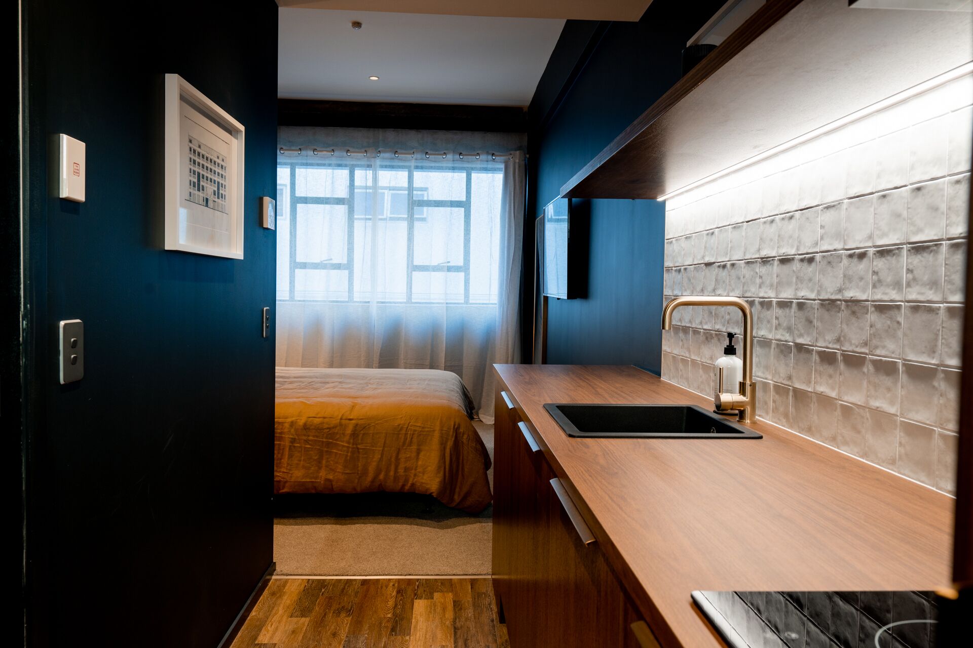 Une chambre d’hôtel moderne avec un bureau en bois, un lit recouvert d’une courtepointe beige, une fenêtre avec des rideaux légers et un mur recouvert de carreaux.