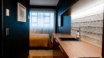 Une chambre d’hôtel moderne avec un bureau en bois, un lit recouvert d’une courtepointe beige, une fenêtre avec des rideaux légers et un mur recouvert de carreaux.