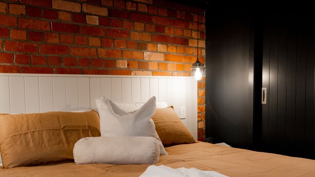 A hotel room with a brick wall, a white headboard, and a bed with brown bedding and pillows. - The Cobbler Hotel (Wellington)