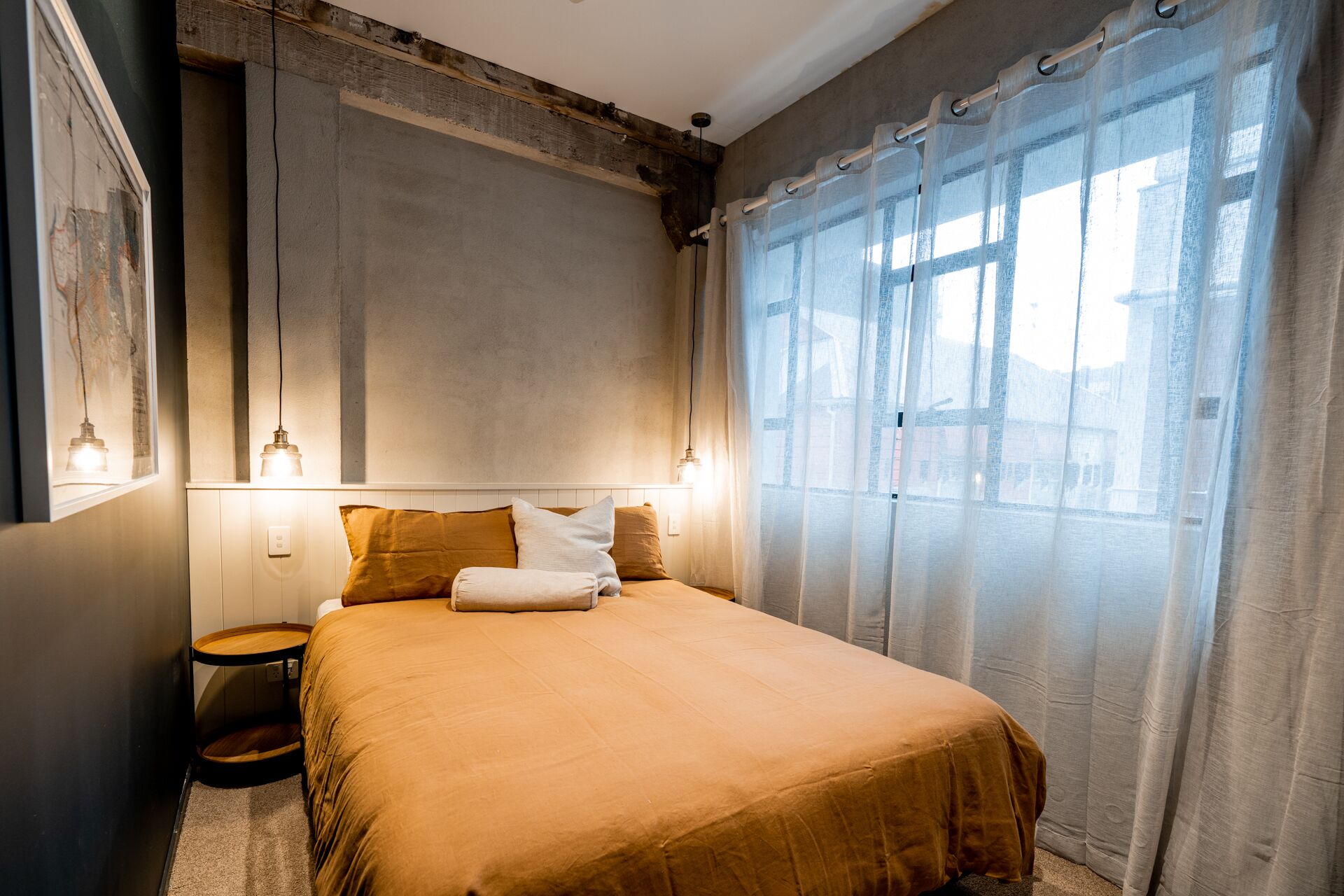 A bedroom with a large bed, a round wooden side table, and a window with sheer curtains.