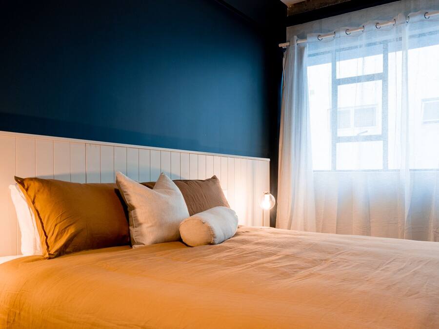 Una cama bien tendida con cabecera blanca, una lámpara de noche y cortinas translúcidas que dejan entrar la luz natural.