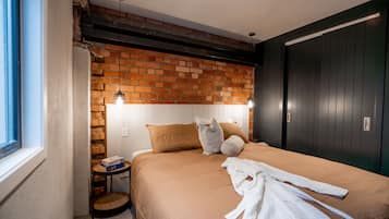 A bedroom with a brick wall, a bed with a brown bedspread, a white robe on the bed, a bedside table with a lamp, and a window.