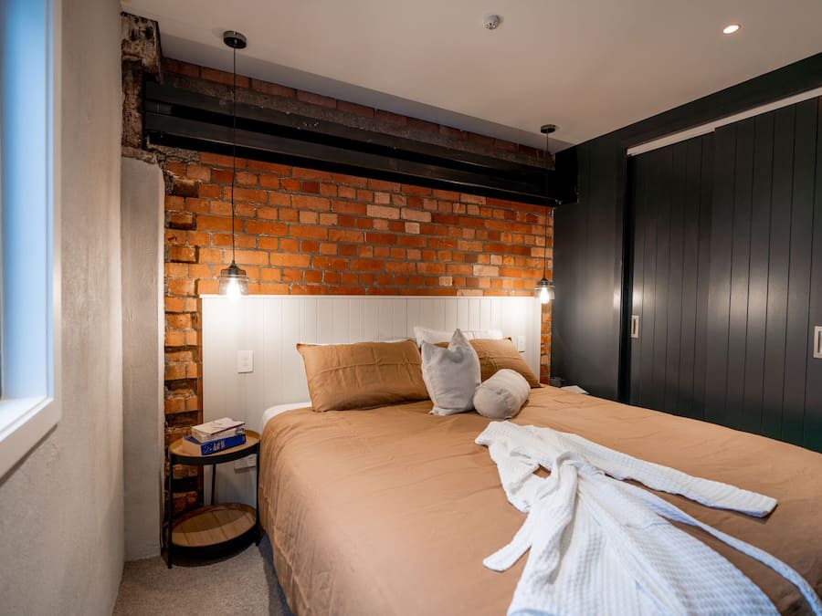 A bedroom with a brick wall, a bed with a brown bedspread, a white robe on the bed, a bedside table with a lamp, and a window.