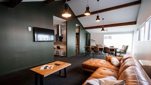 Een moderne woonkamer met een leren bank, een houten salontafel en een aan de muur bevestigde televisie. Er zijn hanglampen en een keuken zichtbaar op de achtergrond.