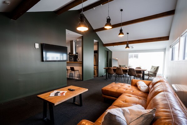 Una sala de estar moderna con un sofá de cuero, una mesa de centro de madera y un televisor montado en la pared. Se observan luces colgantes y una zona de cocina al fondo.