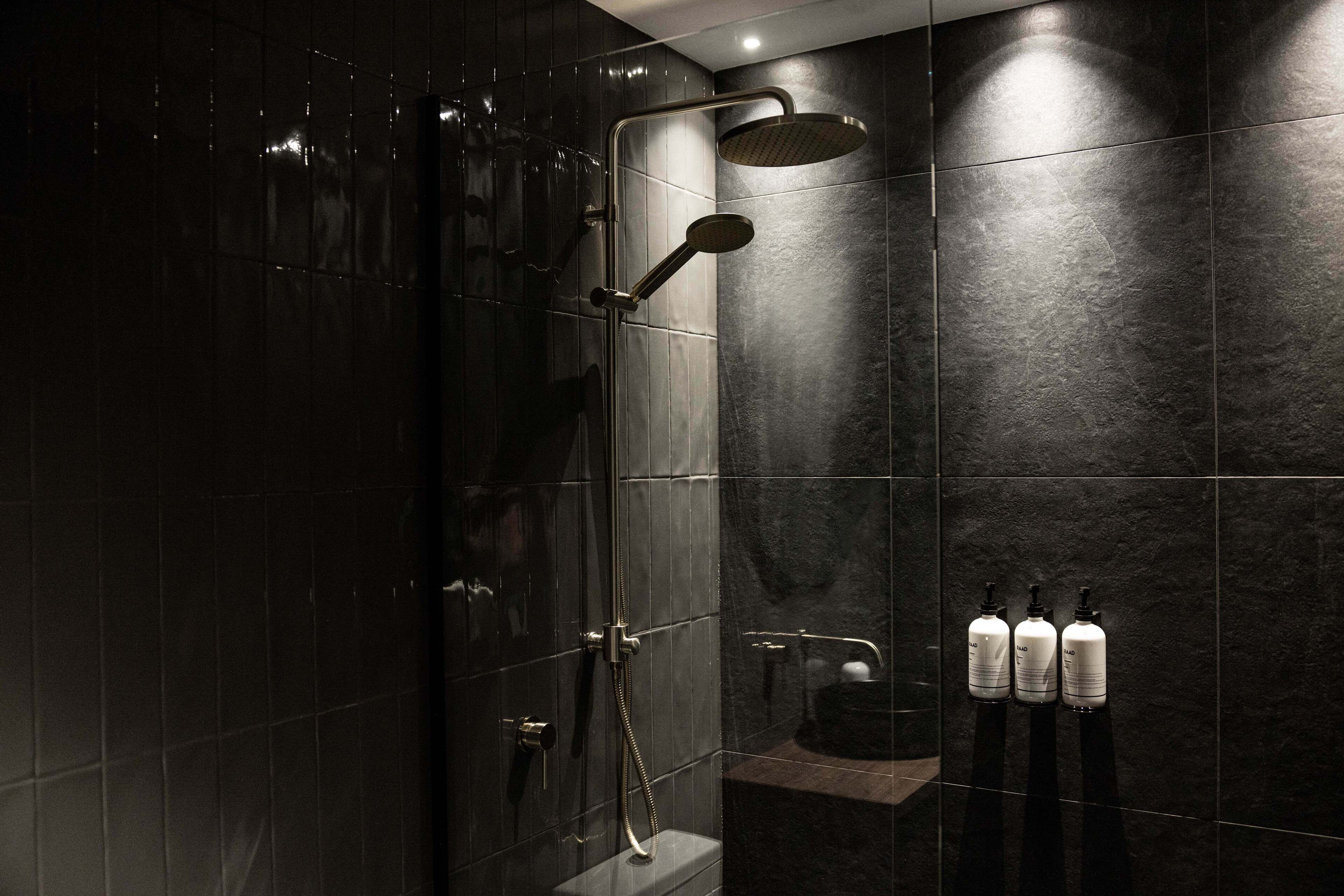 A modern bathroom with dark tiles, a glass-enclosed shower, and three bottles of hand soap on a shelf.