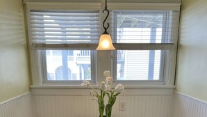 Un comedor con una mesa de madera y bancos blancos, una lámpara colgante y una ventana con persianas.
