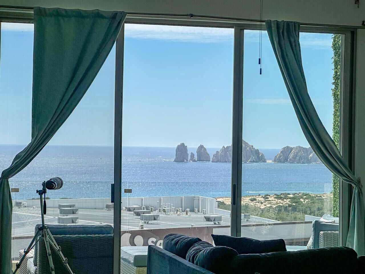 Ein Zimmer mit großem Fenster und Blick auf eine Küstenlandschaft, inklusive Stativ mit Kamera, einer Couch und Ausblick auf das Meer und Felsformationen.