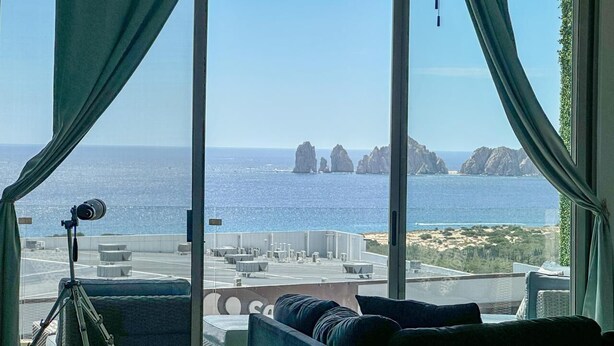Une pièce dotée d’une grande fenêtre donnant sur un paysage côtier, avec un trépied muni d’un appareil photo, un canapé et une vue sur la mer et les formations rocheuses.