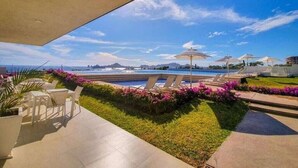 Un espace aménagé au bord de la piscine, avec des chaises longues et des parasols, une vue sur l’océan et un jardin paysager agrémenté de fleurs roses.